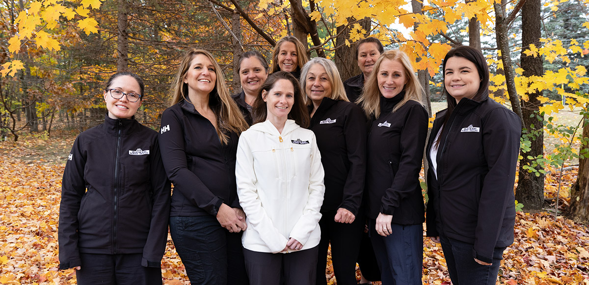 The Williston Road Family Dental team standing together wearing all black except for woman in front. Group is standing outside with fall foliage in the background.